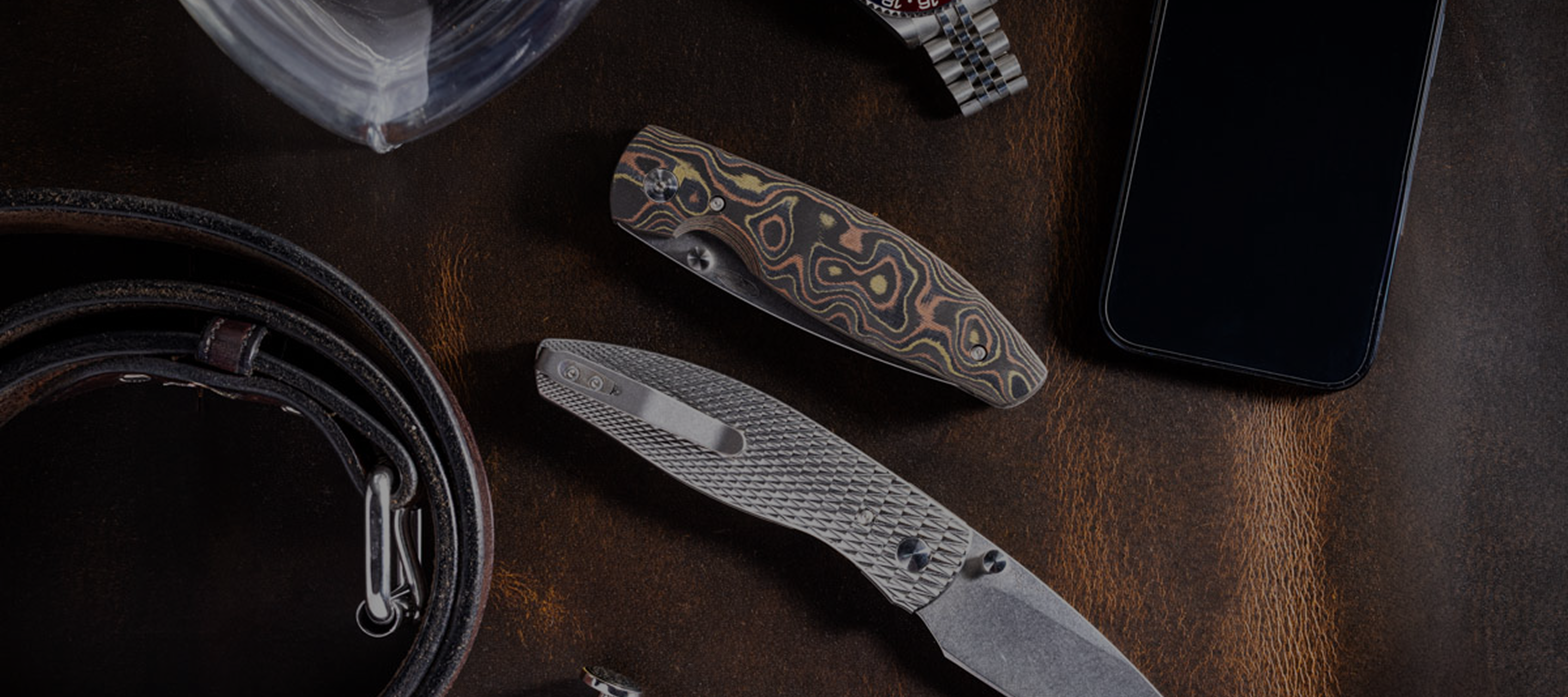 TRM knives on a wooden surface with a phone and belt in the background